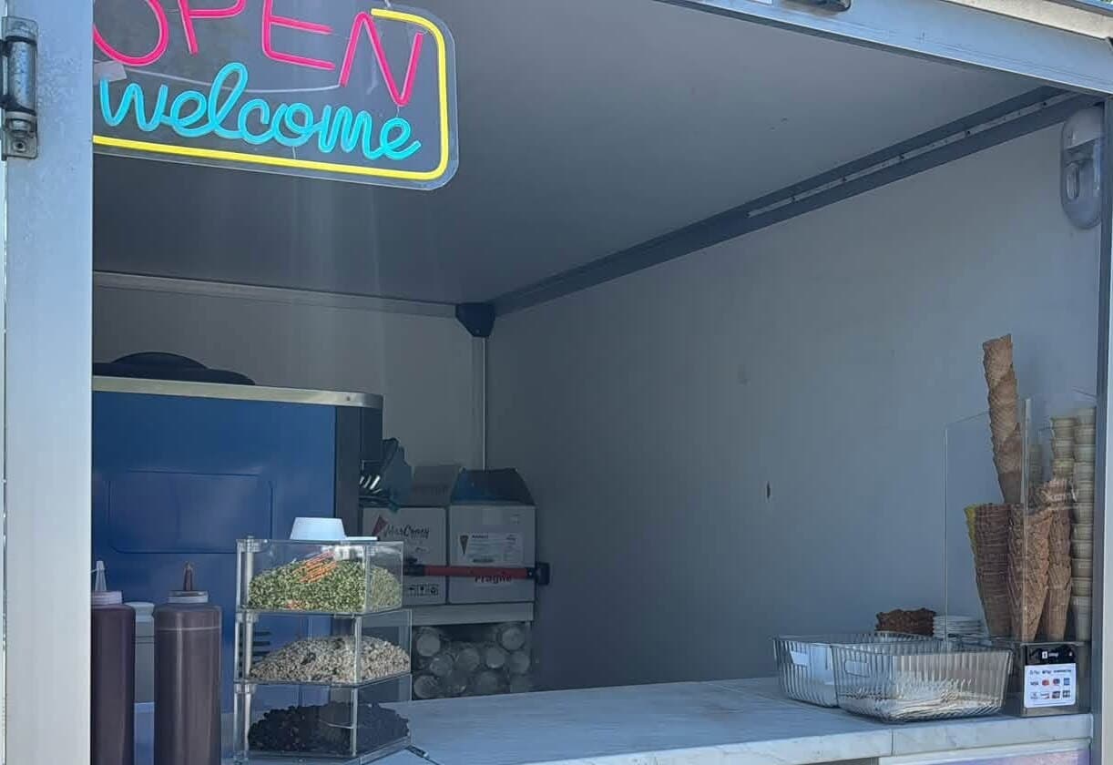 Blick in Mondys geöffneten Verkaufsanhänger: OPEN-welcome-Schild, Softeis-Maschine, Topping-Station, Ausgabe-Theke und die gestapelten Waffeltüten